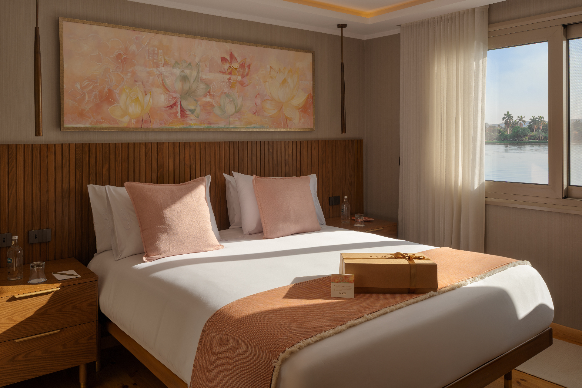 Luxury suite aboard Cleo Dahabiya with king bed, wood-panelled headboard, lotus artwork and a large window overlooking the Nile and palm trees